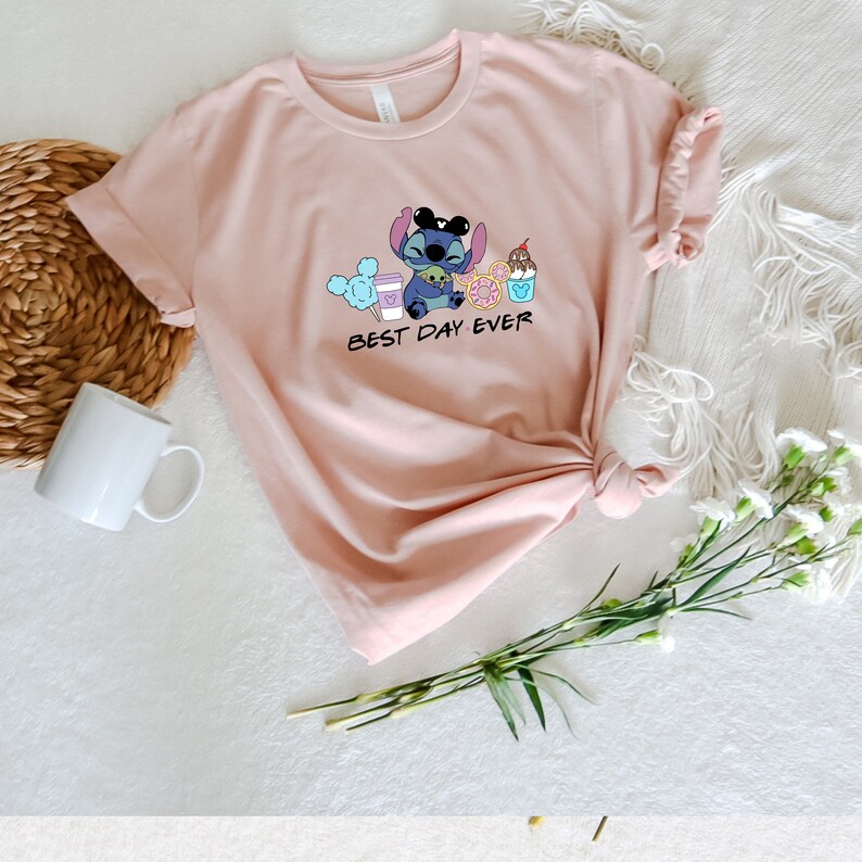 May include: A pink t-shirt with a blue cartoon character holding a cup of coffee, a donut, a Mickey Mouse ice cream cone, and a cotton candy. The text "BEST DAY EVER" is printed below the character.