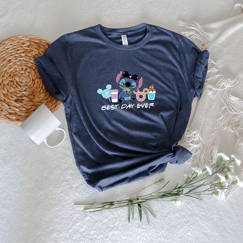 May include: A dark blue t-shirt with a cartoon Stitch character wearing a Mickey Mouse headband and holding a coffee cup, a donut, and a Mickey Mouse ice cream cone. The text "BEST. DAY. EVER." is printed below the character.