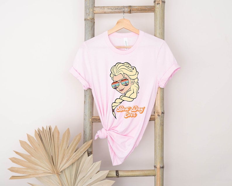 Puede incluir: Camiseta rosa con una imagen de dibujos animados de una mujer con cabello rubio usando gafas de sol y el texto "Best Day Ever".