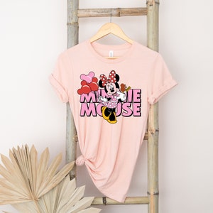 May include: Pink t-shirt with a graphic of Minnie Mouse holding red and pink balloons. The text "Minnie Mouse" is printed in pink letters.