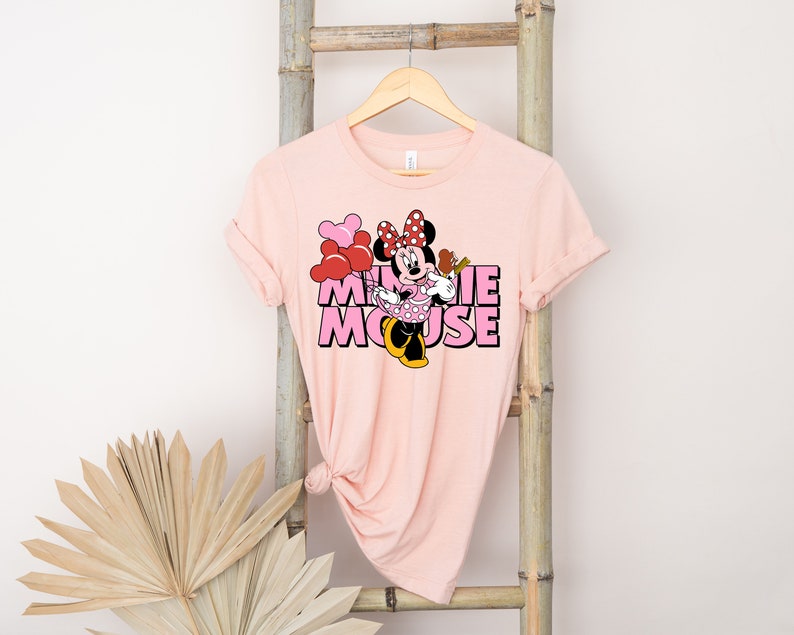 May include: Pink t-shirt with a graphic of Minnie Mouse holding red and pink balloons. The text "Minnie Mouse" is printed in pink letters.