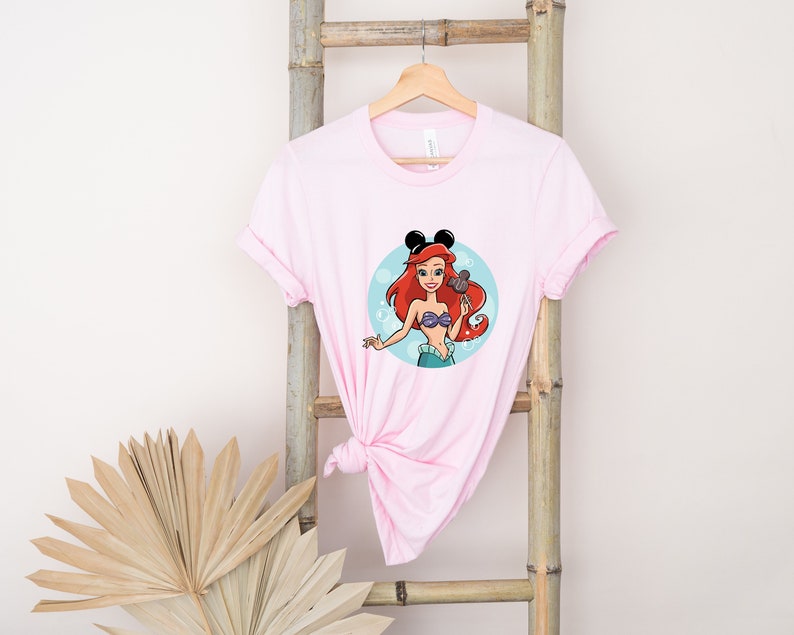 May include: Pink t-shirt with a cartoon image of Ariel from The Little Mermaid wearing Mickey Mouse ears and holding a Mickey Mouse ice cream bar. The image is in a circle with blue bubbles around it.