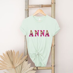 May include: A light green t-shirt with the name "ANNA" printed in pink letters. Each letter is filled with an image of a cartoon girl with brown hair.