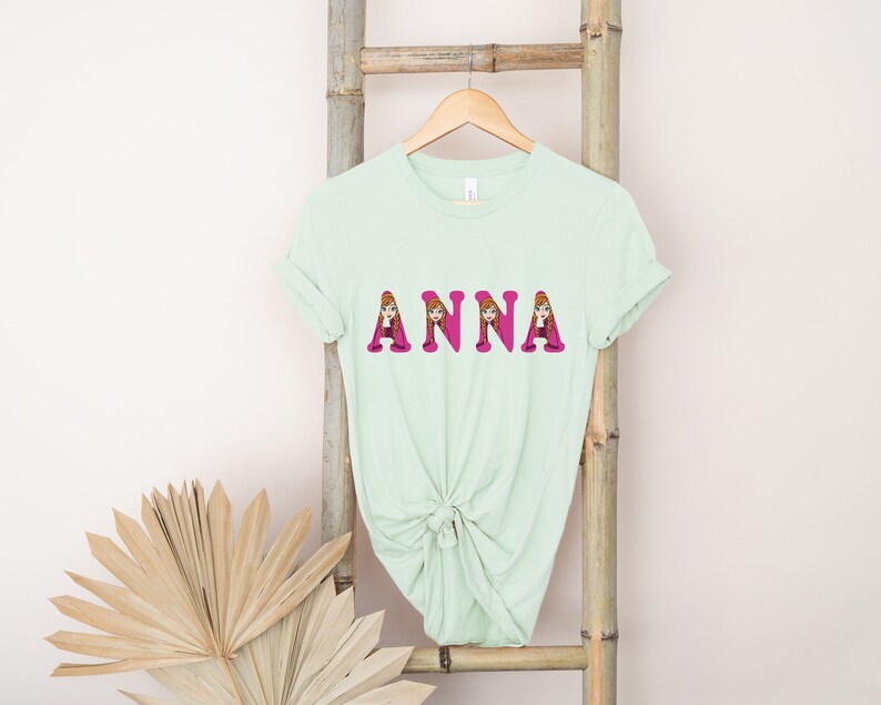May include: A light green t-shirt with the name "ANNA" printed in pink letters. Each letter is filled with an image of a cartoon girl with brown hair.