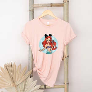 May include: Pink t-shirt with a cartoon image of Ariel from The Little Mermaid wearing Mickey Mouse ears. The image is surrounded by blue bubbles.