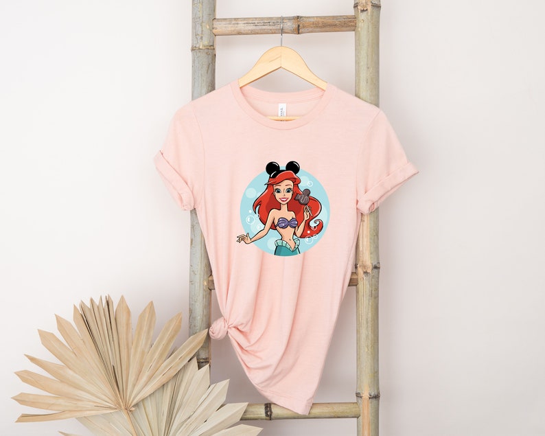 May include: Pink t-shirt with a cartoon image of Ariel from The Little Mermaid wearing Mickey Mouse ears. The image is surrounded by blue bubbles.