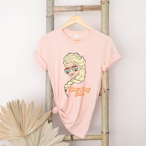 Puede incluir: Una camiseta rosa con una imagen de dibujos animados de una mujer rubia con gafas de sol y el texto "Best Day Ever".