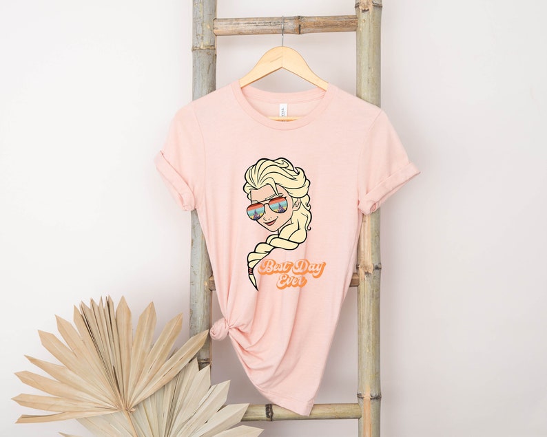 Puede incluir: Una camiseta rosa con una imagen de dibujos animados de una mujer rubia con gafas de sol y el texto "Best Day Ever".