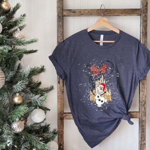 May include: A dark blue t-shirt with a white and gold illustration of Olaf the snowman from Frozen wearing a Santa hat in front of a castle with a red bow on top. The castle is surrounded by white glitter.