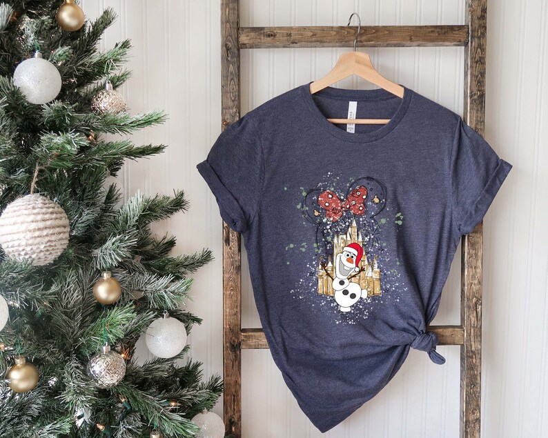 May include: A dark blue t-shirt with a white and gold illustration of Olaf the snowman from Frozen wearing a Santa hat in front of a castle with a red bow on top. The castle is surrounded by white glitter.