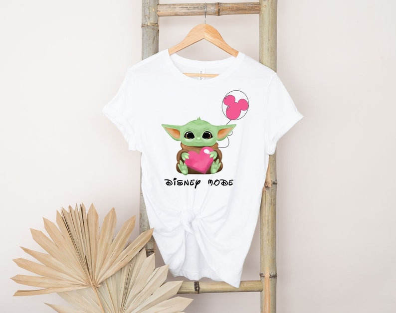 May include: White t-shirt featuring a green Baby Yoda character holding a pink heart and a pink Mickey Mouse balloon. The text "DISNEY MODE" is printed below the character.