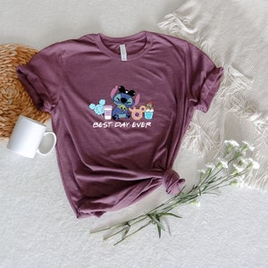 May include: A purple t-shirt with a cartoon Stitch character holding a coffee cup and a donut. The text "BEST DAY EVER" is printed below the character.