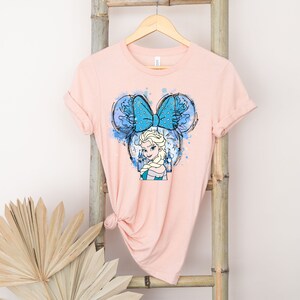 May include: A pink t-shirt with a blue and white watercolor design of Elsa from Frozen wearing a blue bow with white polka dots. The design is in the shape of Mickey Mouse ears.