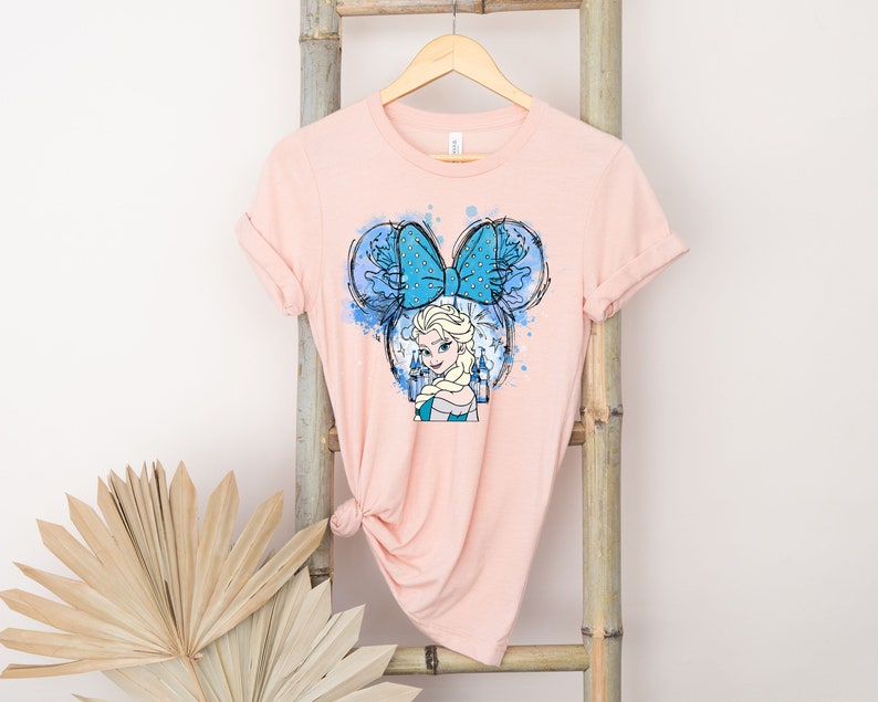 May include: A pink t-shirt with a blue and white watercolor design of Elsa from Frozen wearing a blue bow with white polka dots. The design is in the shape of Mickey Mouse ears.