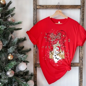 May include: Red t-shirt with a white and gold graphic of Olaf the snowman from Frozen standing in front of a castle with Mickey Mouse balloons. The graphic has a splatter paint effect.