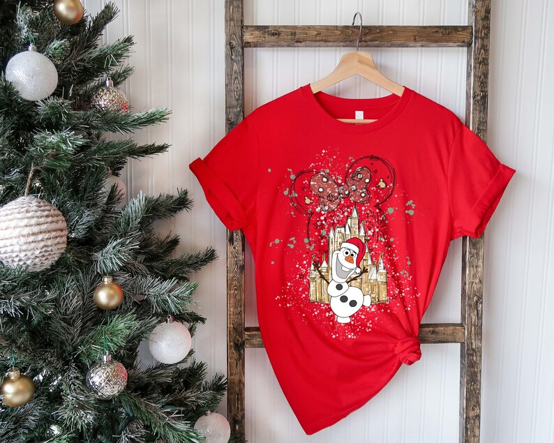 May include: Red t-shirt with a white and gold graphic of Olaf the snowman from Frozen standing in front of a castle with Mickey Mouse balloons. The graphic has a splatter paint effect.