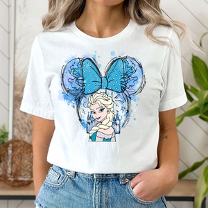 May include: White t-shirt with a blue and white watercolor design of a Disney princess with a blue bow. The princess is wearing a blue dress and has blonde hair. The design is in the shape of Mickey Mouse ears.