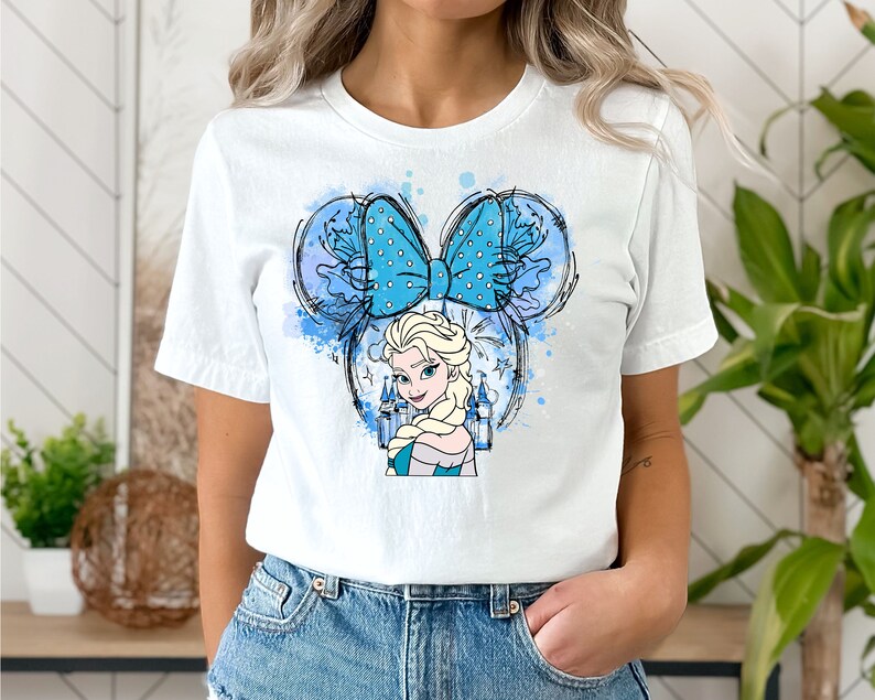 May include: White t-shirt with a blue and white watercolor design of a Disney princess with a blue bow. The princess is wearing a blue dress and has blonde hair. The design is in the shape of Mickey Mouse ears.