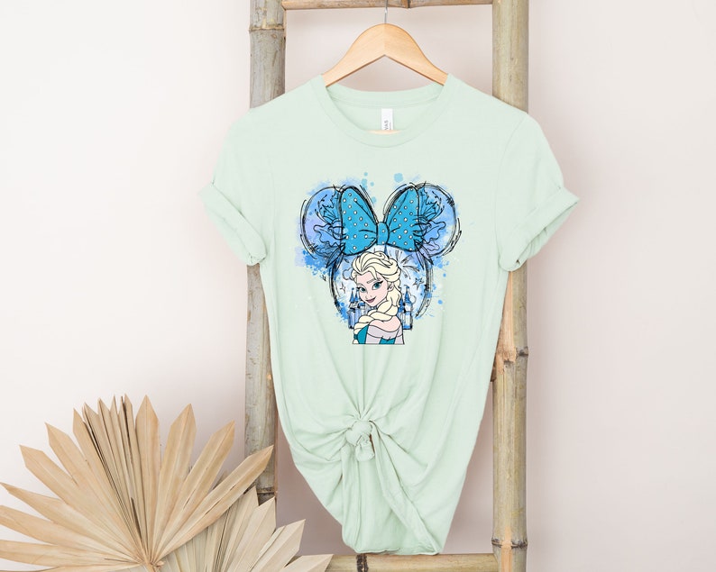 May include: A light green t-shirt with a blue and white illustration of a Disney princess with a blue bow and a castle in the background.