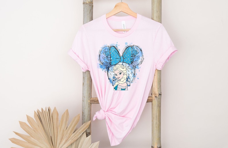 May include: Pink t-shirt with a watercolor illustration of Elsa from Frozen wearing a blue bow and Mickey Mouse ears.