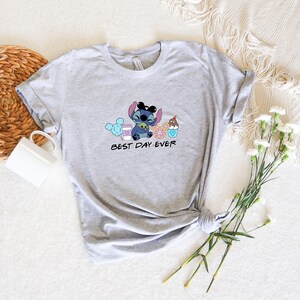 May include: Gray t-shirt with a blue Stitch character wearing Mickey Mouse ears and holding a green alien. Stitch is surrounded by cartoon images of cotton candy, a coffee cup, a donut, and a Mickey Mouse ice cream cone. The text "BEST DAY EVER" is printed below Stitch.
