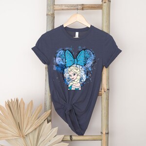 May include: A dark blue t-shirt with a watercolor design featuring a cartoon image of Elsa from Frozen wearing a blue bow with white polka dots. The design is reminiscent of Mickey Mouse ears.