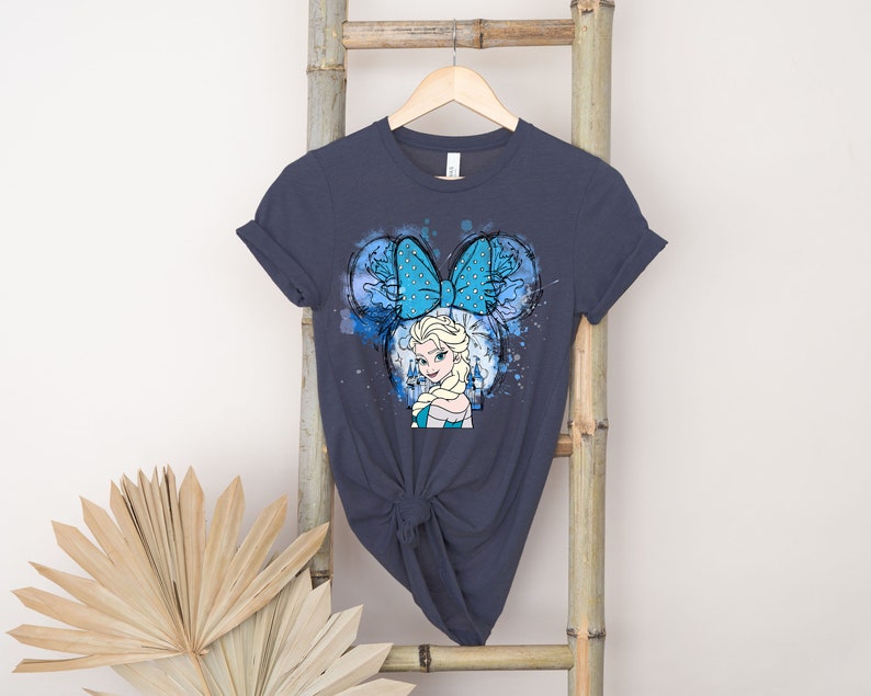 May include: A dark blue t-shirt with a watercolor design featuring a cartoon image of Elsa from Frozen wearing a blue bow with white polka dots. The design is reminiscent of Mickey Mouse ears.