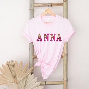 May include: Pink t-shirt with the name "ANNA" spelled out in pink letters. Each letter is decorated with a cartoon image of Anna from the movie Frozen.