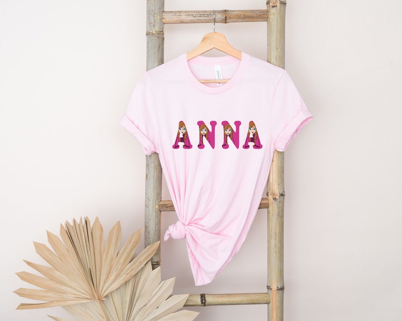 May include: Pink t-shirt with the name "ANNA" spelled out in pink letters. Each letter is decorated with a cartoon image of Anna from the movie Frozen.