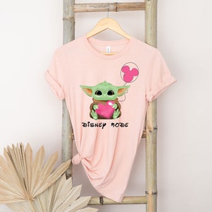 May include: Pink t-shirt featuring a green Baby Yoda character holding a pink heart and a pink Mickey Mouse balloon. The text "DISNEY MODE" is printed on the shirt.
