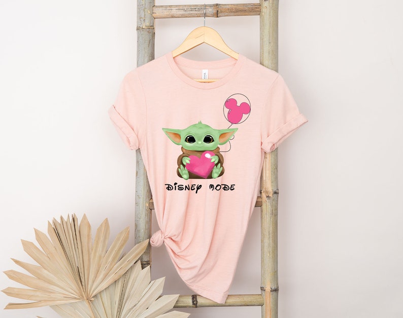 May include: Pink t-shirt featuring a green Baby Yoda character holding a pink heart and a pink Mickey Mouse balloon. The text "DISNEY MODE" is printed on the shirt.