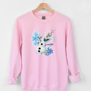 May include: Pink sweatshirt with a graphic of Olaf from Frozen. Olaf is smiling and has his arms outstretched. The background is blue and white with snowflakes.