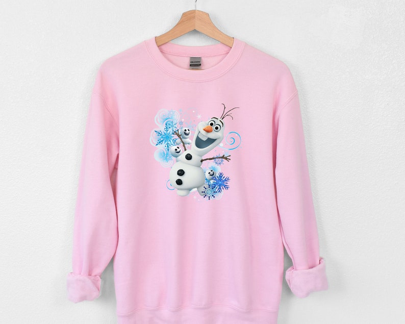 May include: Pink sweatshirt with a graphic of Olaf from Frozen. Olaf is smiling and has his arms outstretched. The background is blue and white with snowflakes.