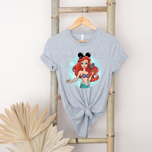 May include: A gray t-shirt with a cartoon image of Ariel from The Little Mermaid wearing Mickey Mouse ears and holding a Mickey Mouse ice cream bar. The image is surrounded by blue bubbles.