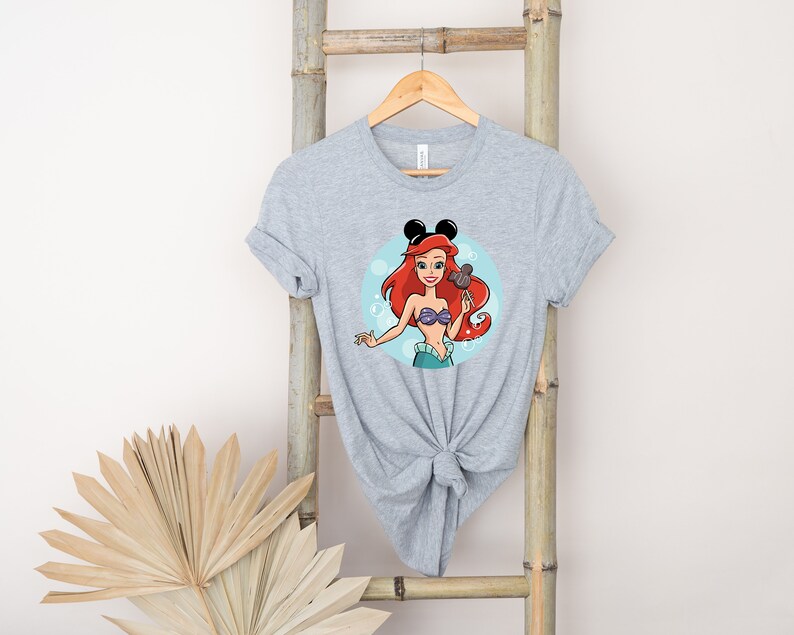 May include: A gray t-shirt with a cartoon image of Ariel from The Little Mermaid wearing Mickey Mouse ears and holding a Mickey Mouse ice cream bar. The image is surrounded by blue bubbles.