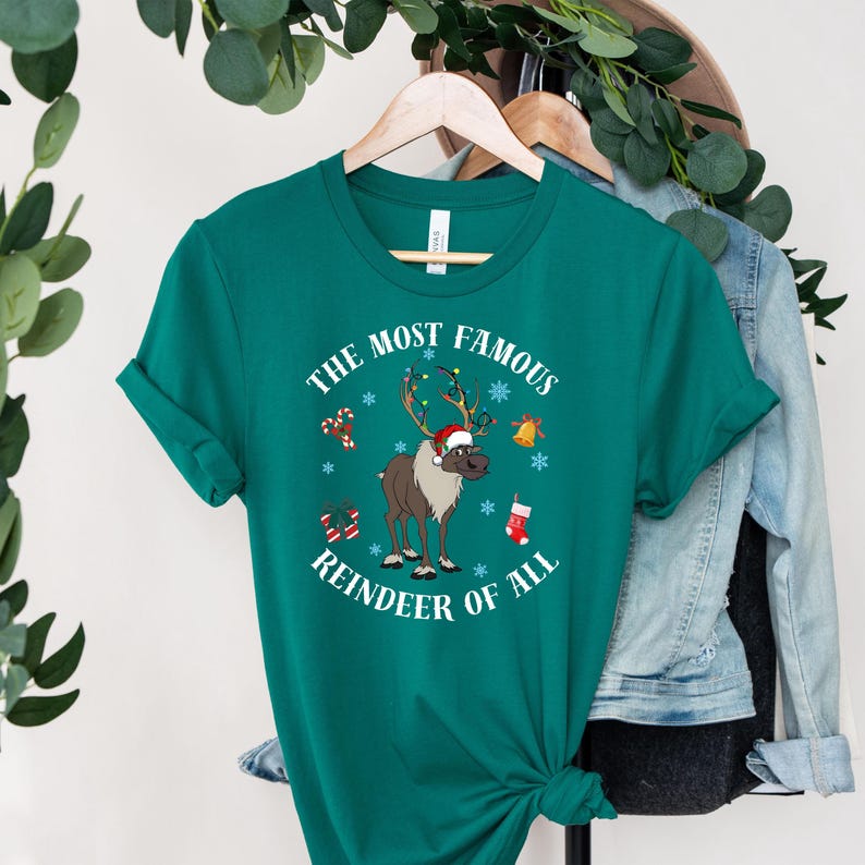 Pu&ograve; includere: Una maglietta verde con una grafica bianca di una renna con un cappello di Babbo Natale rosso e il testo "The Most Famous Reindeer of All".