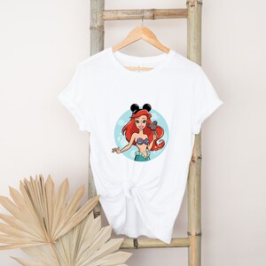 May include: White t-shirt with a cartoon image of Ariel from The Little Mermaid wearing Mickey Mouse ears and holding a seashell. The image is in a circle with blue bubbles around it.