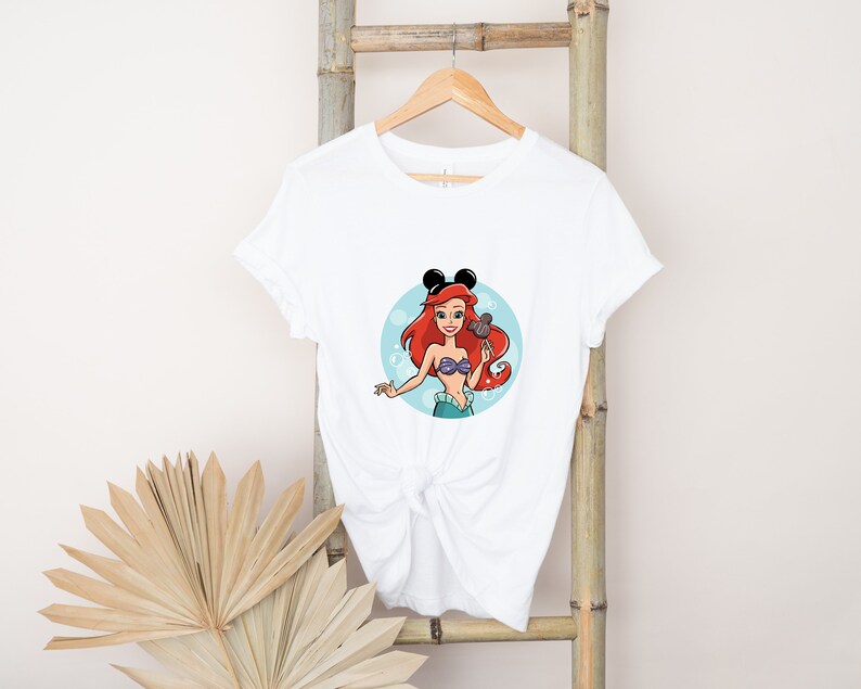 May include: White t-shirt with a cartoon image of Ariel from The Little Mermaid wearing Mickey Mouse ears and holding a seashell. The image is in a circle with blue bubbles around it.