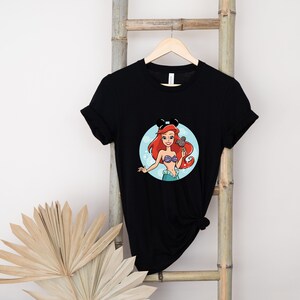 May include: Black t-shirt with a graphic of Ariel from The Little Mermaid wearing Mickey Mouse ears and holding a Mickey Mouse ice cream bar. The graphic is on a light blue circle with white bubbles.