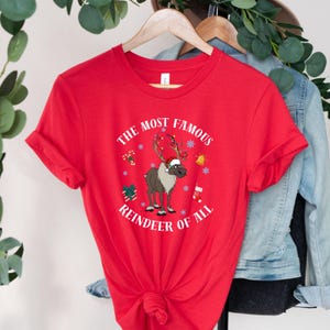 Pu&ograve; includere: Maglietta rossa con una grafica bianca di una renna con un cappello di Babbo Natale e il testo "The Most Famous Reindeer of All".
