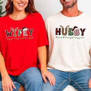 May include: Red and cream Christmas t-shirts. The red shirt says "WIFEY" with reindeer antlers and a Santa hat. The cream shirt says "HUBBY" with antlers and a Santa hat. Both shirts have a string of Christmas lights.