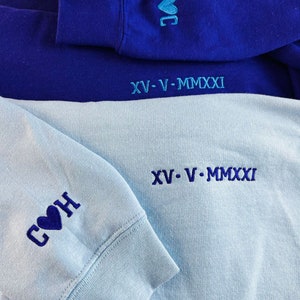 Peut inclure: Deux sweats &agrave; capuche, l'un bleu et l'autre bleu clair, avec des c&oelig;urs brod&eacute;s et le texte "COH" et "HOC" sur les manches. La date "XV-V-MMXXI" est brod&eacute;e au bas de chaque sweat-shirt.