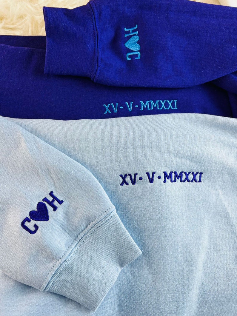 Peut inclure: Deux sweats &agrave; capuche, l'un bleu et l'autre bleu clair, avec des c&oelig;urs brod&eacute;s et le texte "COH" et "HOC" sur les manches. La date "XV-V-MMXXI" est brod&eacute;e au bas de chaque sweat-shirt.