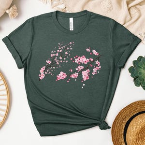 Puede incluir: Una camiseta verde oscuro con un dise&ntilde;o de flores de cerezo rosa y blanco. La camiseta est&aacute; hecha de un material suave y c&oacute;modo.