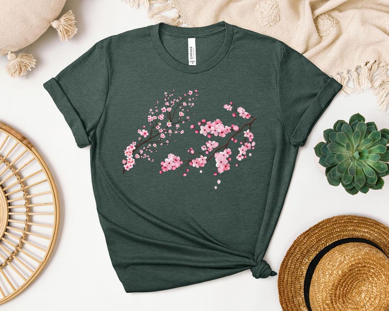 Puede incluir: Una camiseta verde oscuro con un dise&ntilde;o de flores de cerezo rosa y blanco. La camiseta est&aacute; hecha de un material suave y c&oacute;modo.