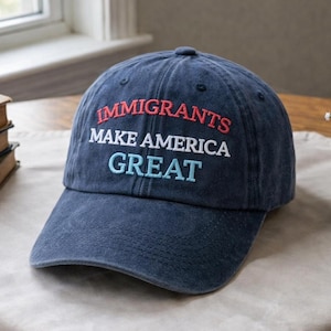 May include: A navy blue baseball cap with the embroidered text "IMMIGRANTS MAKE AMERICA GREAT" in red, white, and light blue. The hat is on a table with books and a small vase of flowers.
