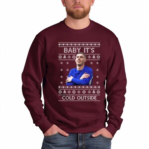 May include: A maroon Christmas sweater with a white snowflake pattern and a picture of a man in a blue jersey. The sweater has the text "Baby It's Cold Outside" printed on it.