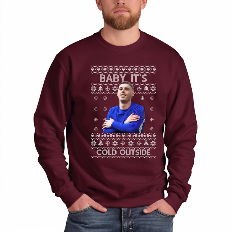 May include: A maroon Christmas sweater with a white snowflake pattern and a picture of a man in a blue jersey. The sweater has the text "Baby It's Cold Outside" printed on it.
