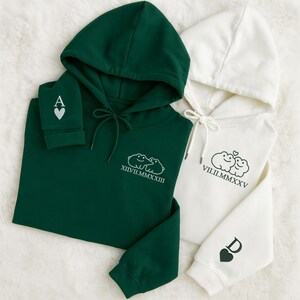 May include: Two hooded sweatshirts, one dark green and one white, are shown. The green sweatshirt features an "A" and a heart on the sleeve, and a cloud graphic with Roman numerals. The white sweatshirt has a "D" and a heart on the sleeve, and a cloud graphic with Roman numerals.