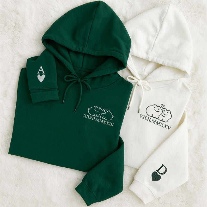 May include: Two hooded sweatshirts, one dark green and one white, are shown. The green sweatshirt features an "A" and a heart on the sleeve, and a cloud graphic with Roman numerals. The white sweatshirt has a "D" and a heart on the sleeve, and a cloud graphic with Roman numerals.